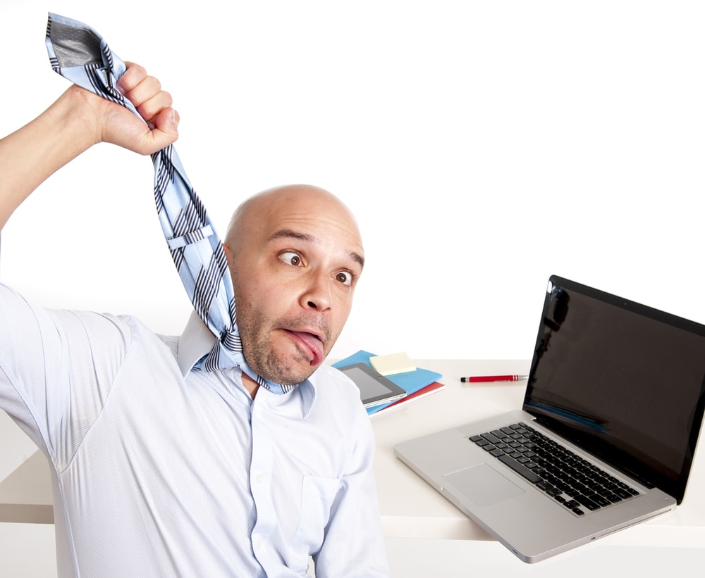 shutterstock_188184989 young bald south american business man choking himself with his own tie in desperation while being in stress , overworked and frustrated while working with computer at office desk