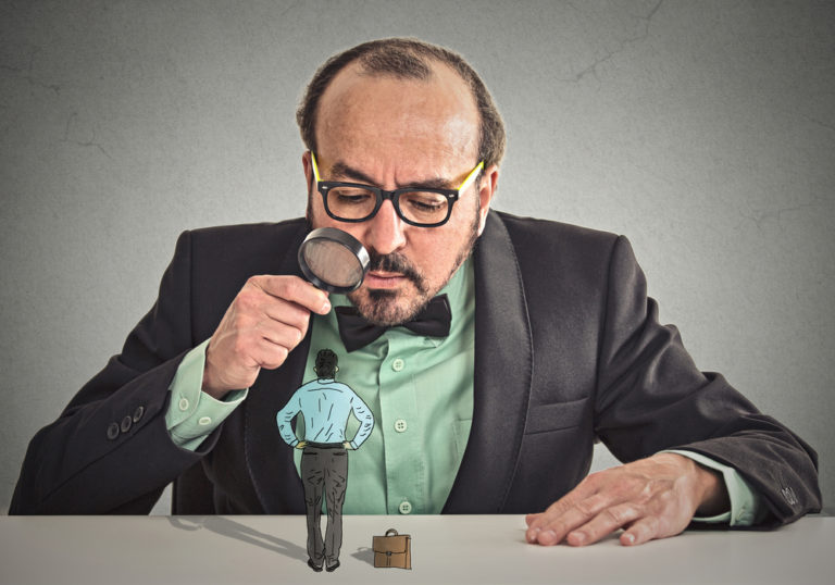 shutterstock_226179493 Curious corporate businessman skeptically meeting looking at small employee standing on table through magnifying glass isolated office grey wall background. Human face expression, attitude, perception