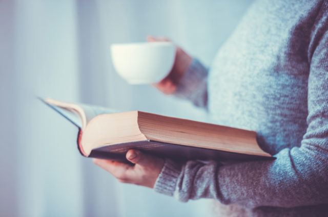 shutterstock_267312578 Young woman reading a book and holding cup of tea or coffee. Toned image
