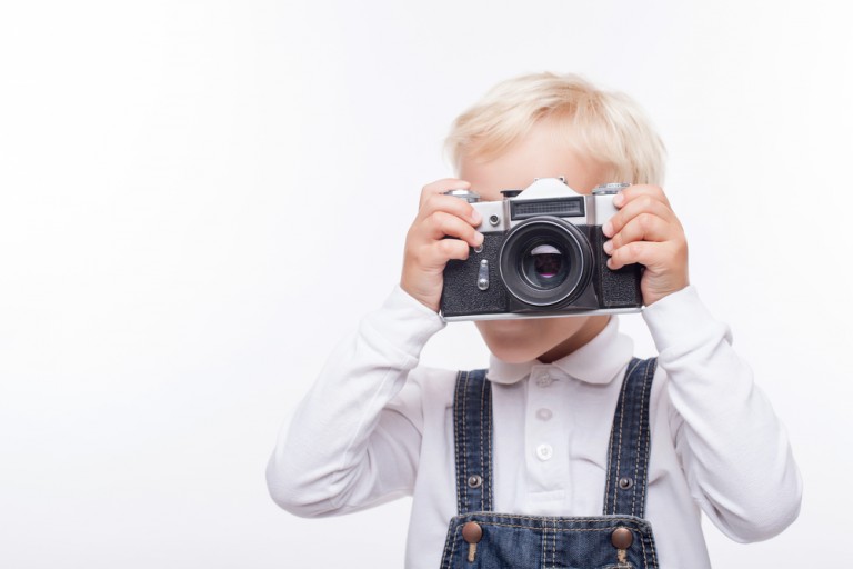 shutterstock_313224770 Cheerful blond boy is standing and holding old camera. He is photographing something with interest. Isolated and copy space in left side
