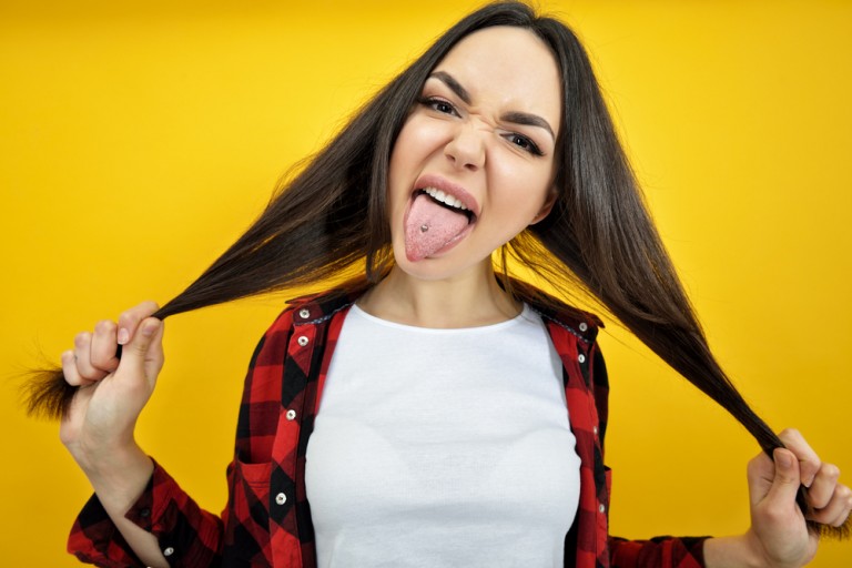 shutterstock_390670672 Hipster girl in checked shirt showing tongue with piercing over yellow background. Impertinent behaviour. Hipsters. Provocation. Aggression. Naughtiness.