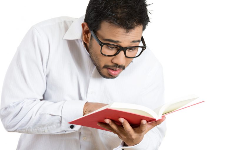 shutterstock_174657788 Closeup portrait of young man, wide opened eyes mouth pointing at a page inside book, shocked surprised by the twists and turn of story, isolated on white background. Human emotion facial expression