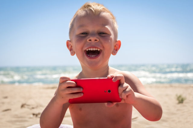 shutterstock_348723281 Cute boy laughing while playing with smart phone at beach near the sea at sunny day